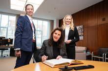 Die Botschafterin sitzt am Besprechungstisch im Präsidentinnenzimmer und trägt sich in der vor ihr liegende Gästebuch ein. Die Vizepräsidentin und die Honorarkonsulin stehen neben ihr. Alle gucken freundlich in die Kamera.