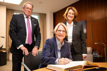 Die Botschafterin sitzt am Konferenztisch im Präsidentinnenzimmer und trägt sich in das Gästebuch ein. Die Präsidentin und der Konsul stehen neben ihr. Alle gucken freundlich in die Kamera.