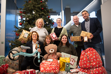 Mehrere Personen knien und stehen nebeneinander vor dem Wunschbaum. Alle halten ein verpacktes Geschenk in ihren Händen und lächeln in die Kamera.