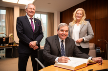 Der Botschafter sitzt am Konferenztisch im Präsidentinnenzimmer und trägt sich in das Gästebuch ein. Hinter ihm stehen die Viezepräsidentin und der Honorarkonsul. Alle gucken freundlich in die Kamera.