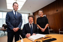 Der Botschafter sitzt am Konferenztisch im Präsidentinnenzimmer und trägt sich in das vor ihm liegende Gästebuch ein. Die Vizepräsidentin und der Honorarkonsul stehen neben ihm. Alle gucken freundlich in die Kamera.