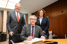 Der Botschafter sitzt am Konferenztisch im Präsidentinnenzimmer und trägt sich in das vor ihm liegende Gästebuch ein. Der Honorarkonsul und die Präsidentin stehen neben ihm. Alle gucken freundlich in die Kamera.