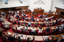 Die Abgeordneten sitzen an ihren Tischen im Plenarsaal und heben ihre Hände zur Abstimmung. Senator:innen und Staatsrät:innen sitzen auf der Regierungsbank links und rechts vom Präsidium.